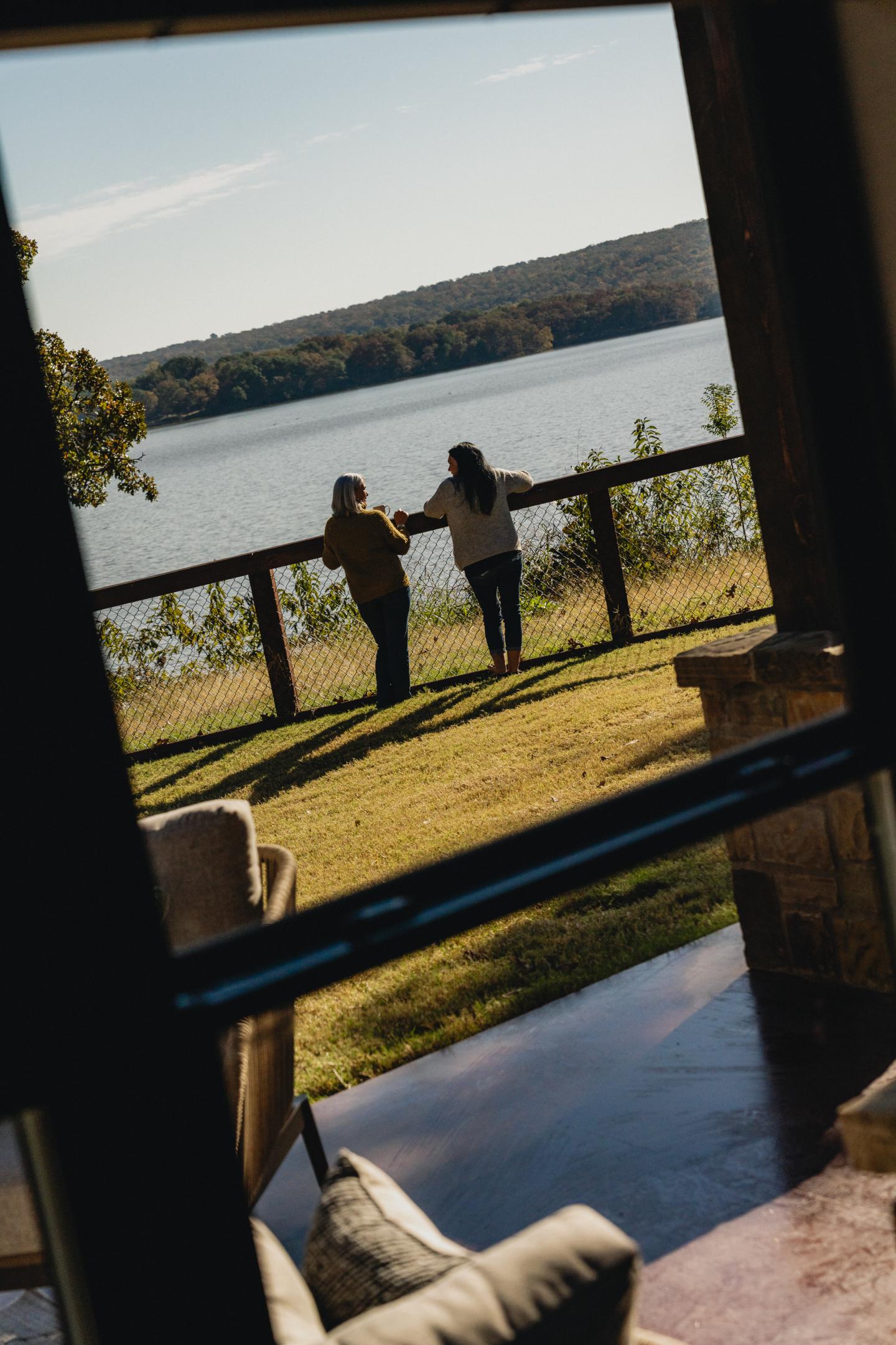 Two people by a lakeside railing, viewed through a window on a sunny day.