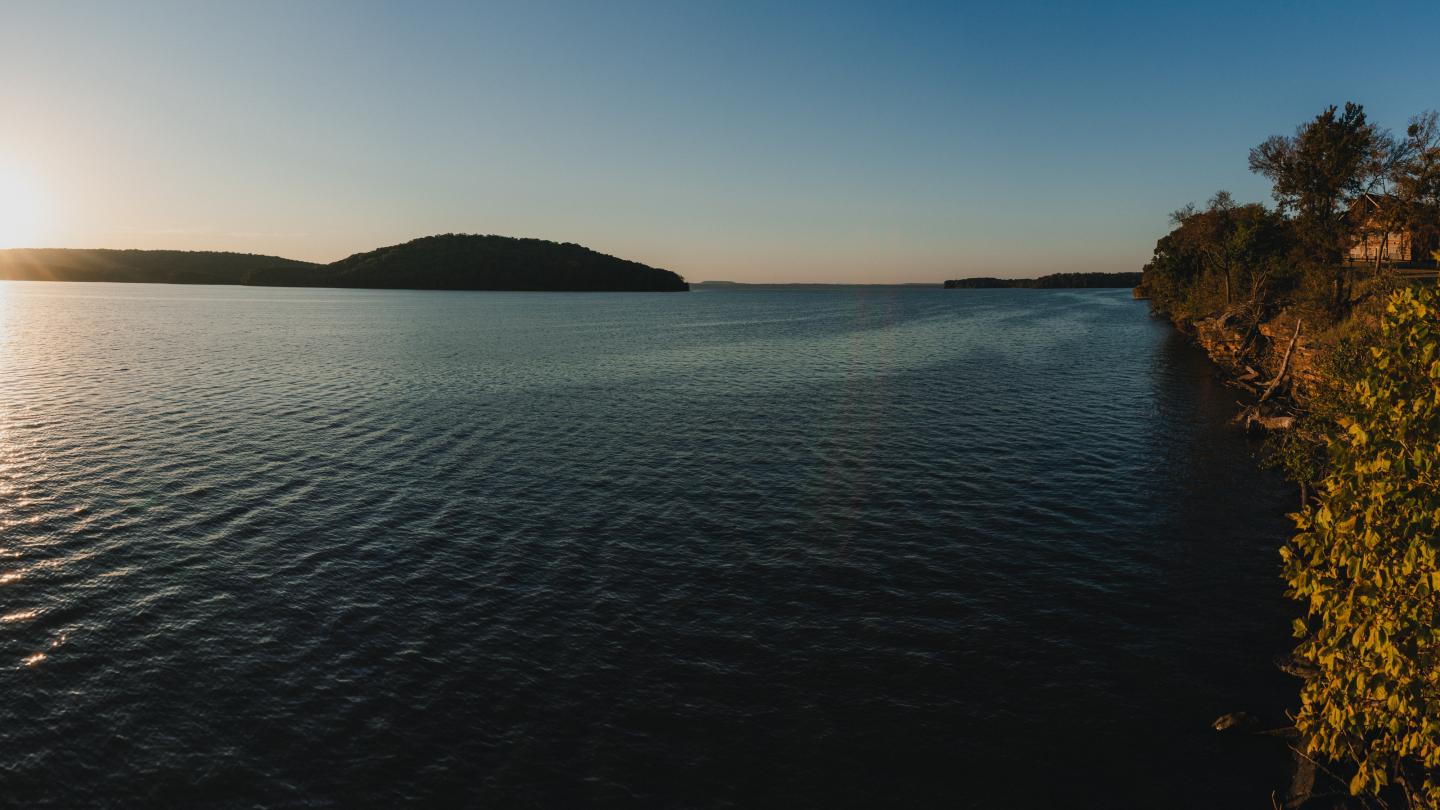 Sunset over a calm lake with distant hills and a tree-lined shore.