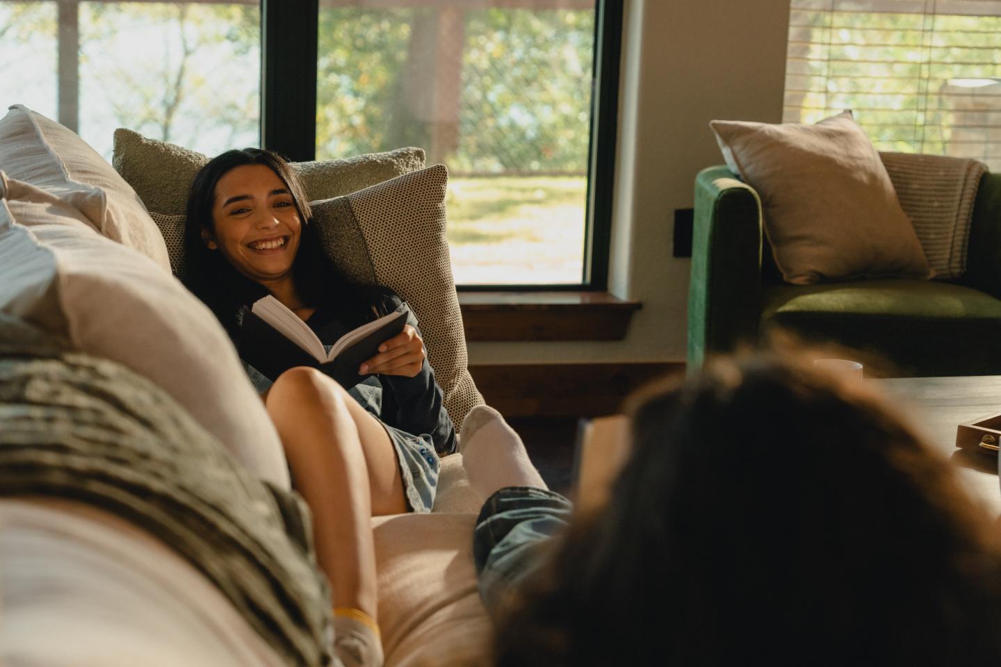 Woman smiling and reading on a couch, sunlit room, relaxed atmosphere.