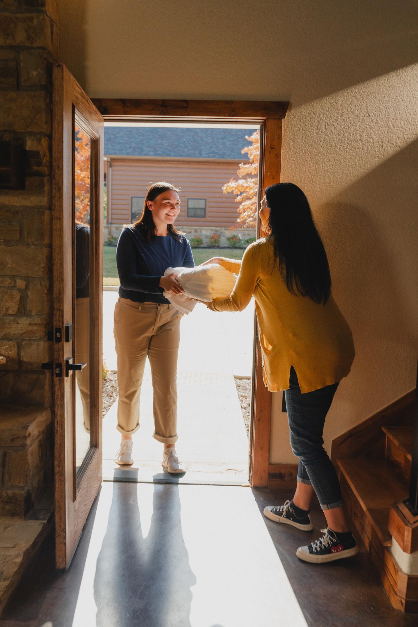 Two women exchange a plate at a sunlit doorway, one inside and one outside.