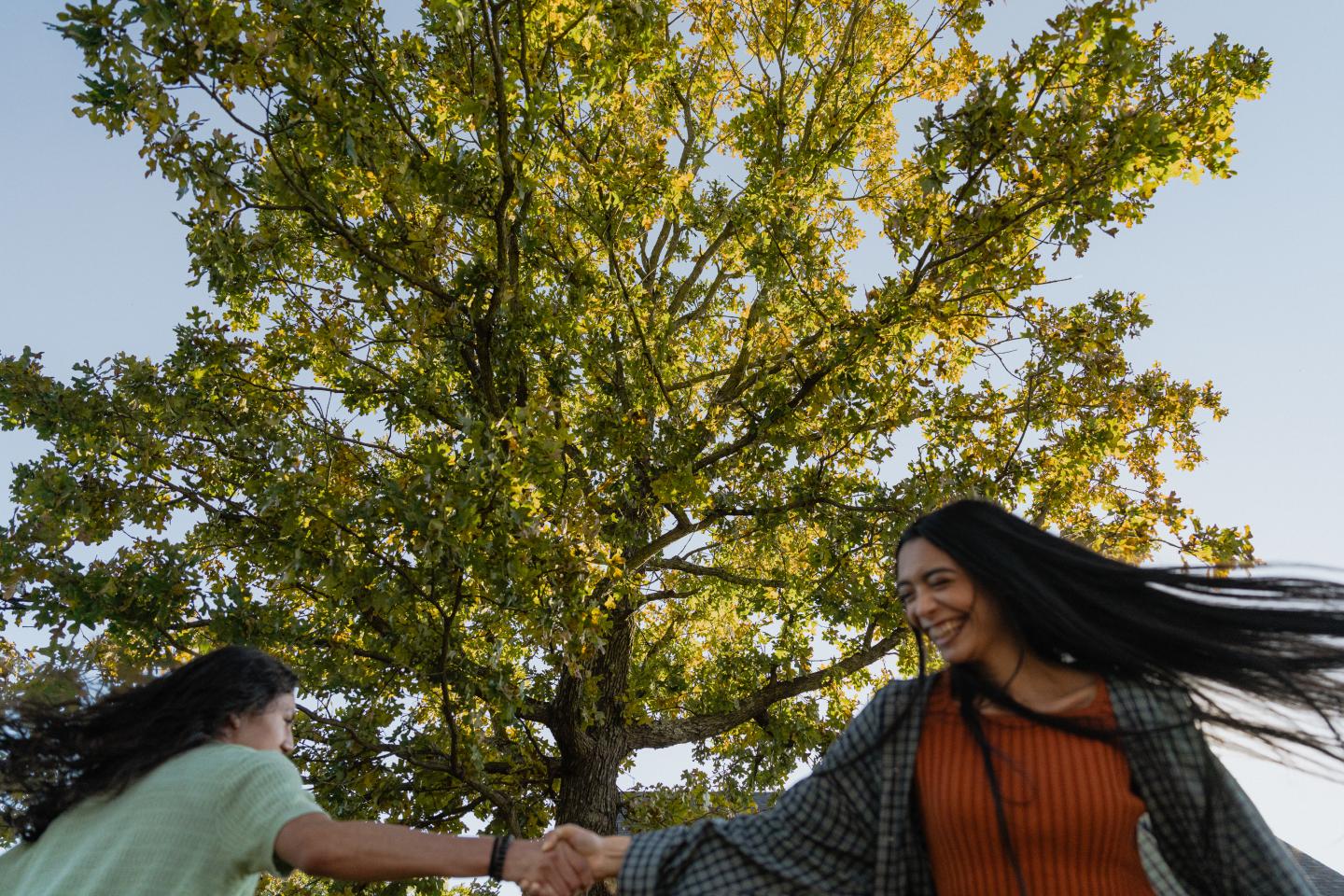 Two people holding hands, smiling in front of a sunlit tree.