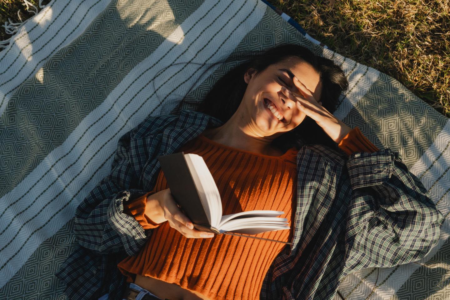 Young woman smiling, lying on a blanket, reading a book in sunlight.