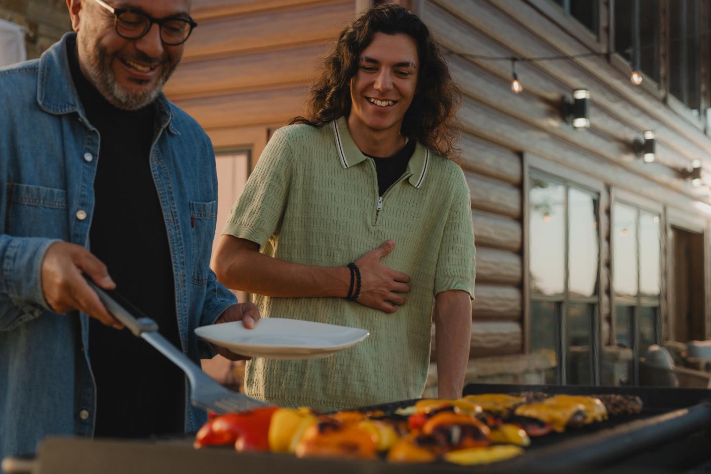 Two people smiling while grilling vegetables outdoors at sunset.