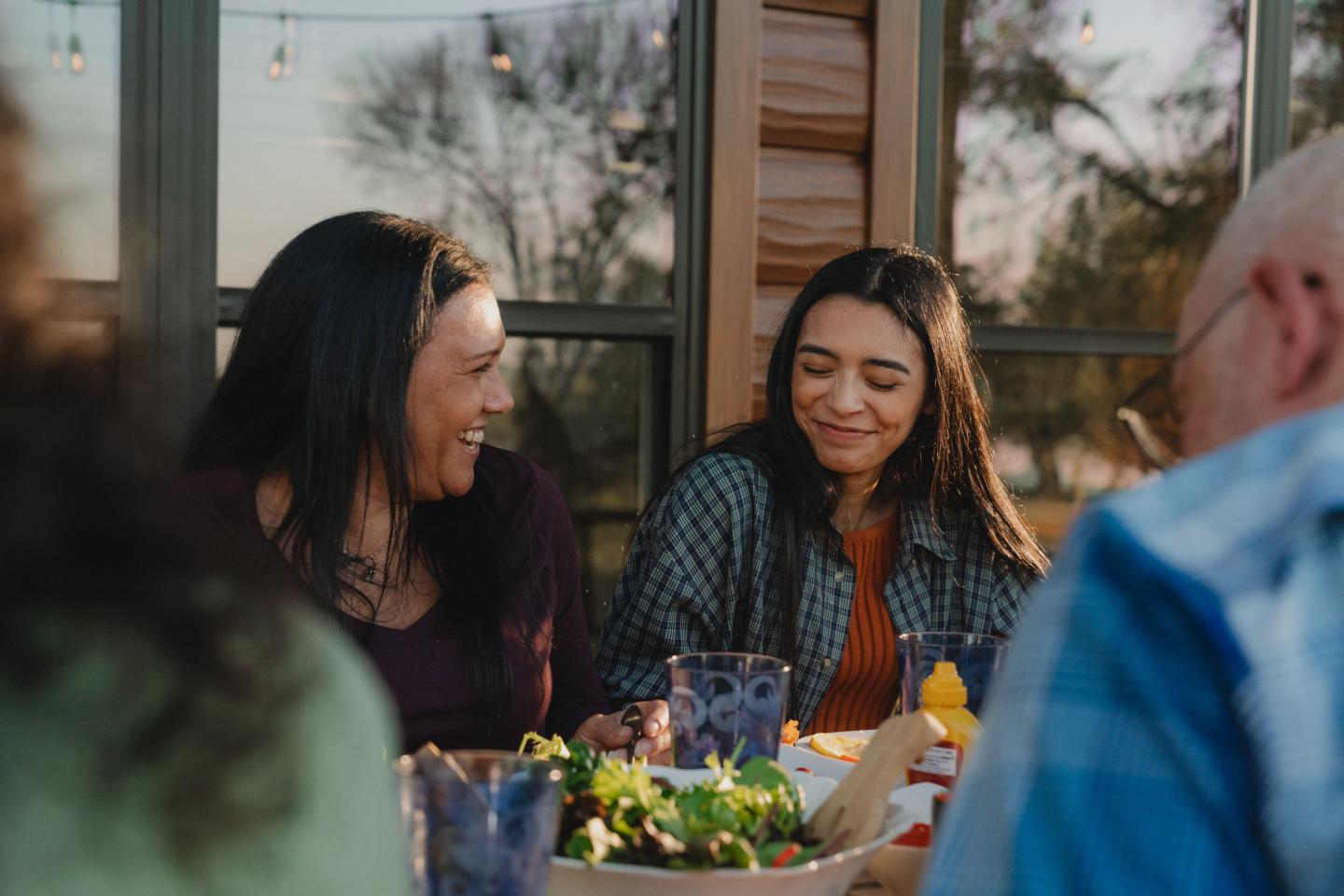 Two women smiling at an outdoor dinner table with blurred figures around them.