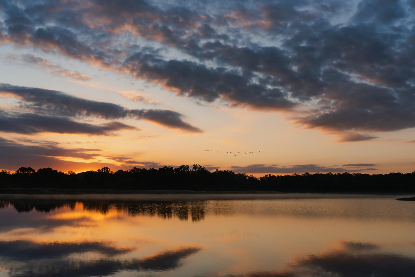 Sunset over a lake with clouds and trees reflecting in the water.