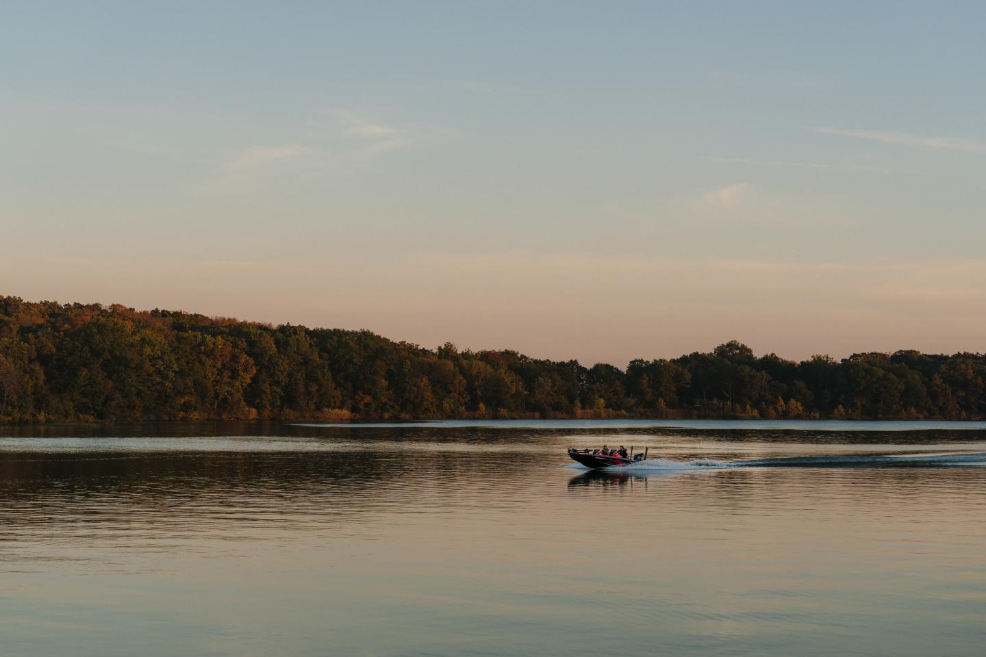 Boat speeding on a calm lake at sunset with trees in the distance.