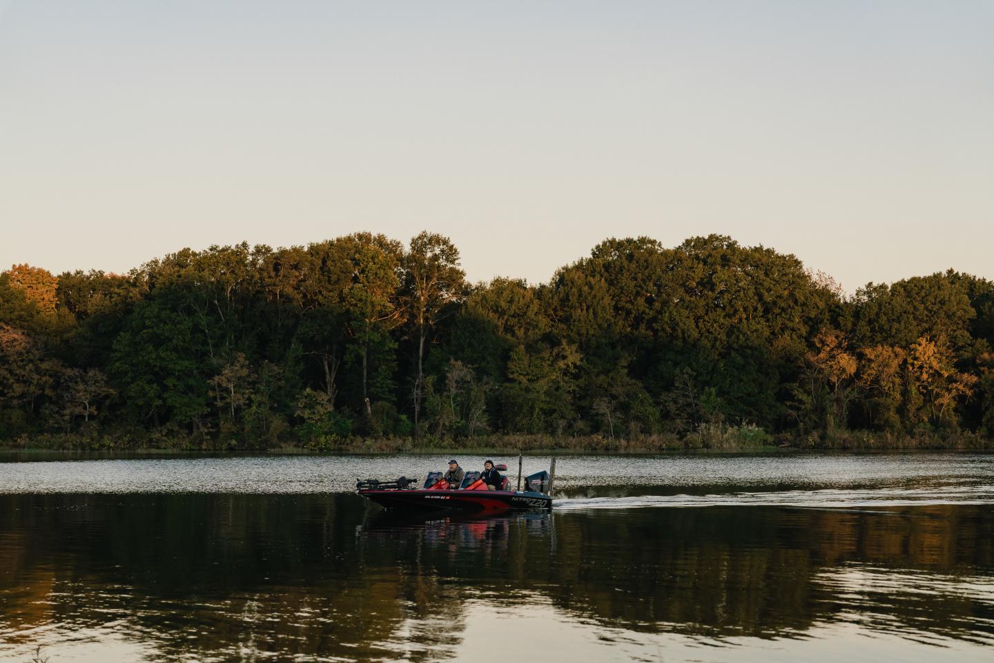 Boat gliding on a calm lake with a wooded shore in the background at sunset.