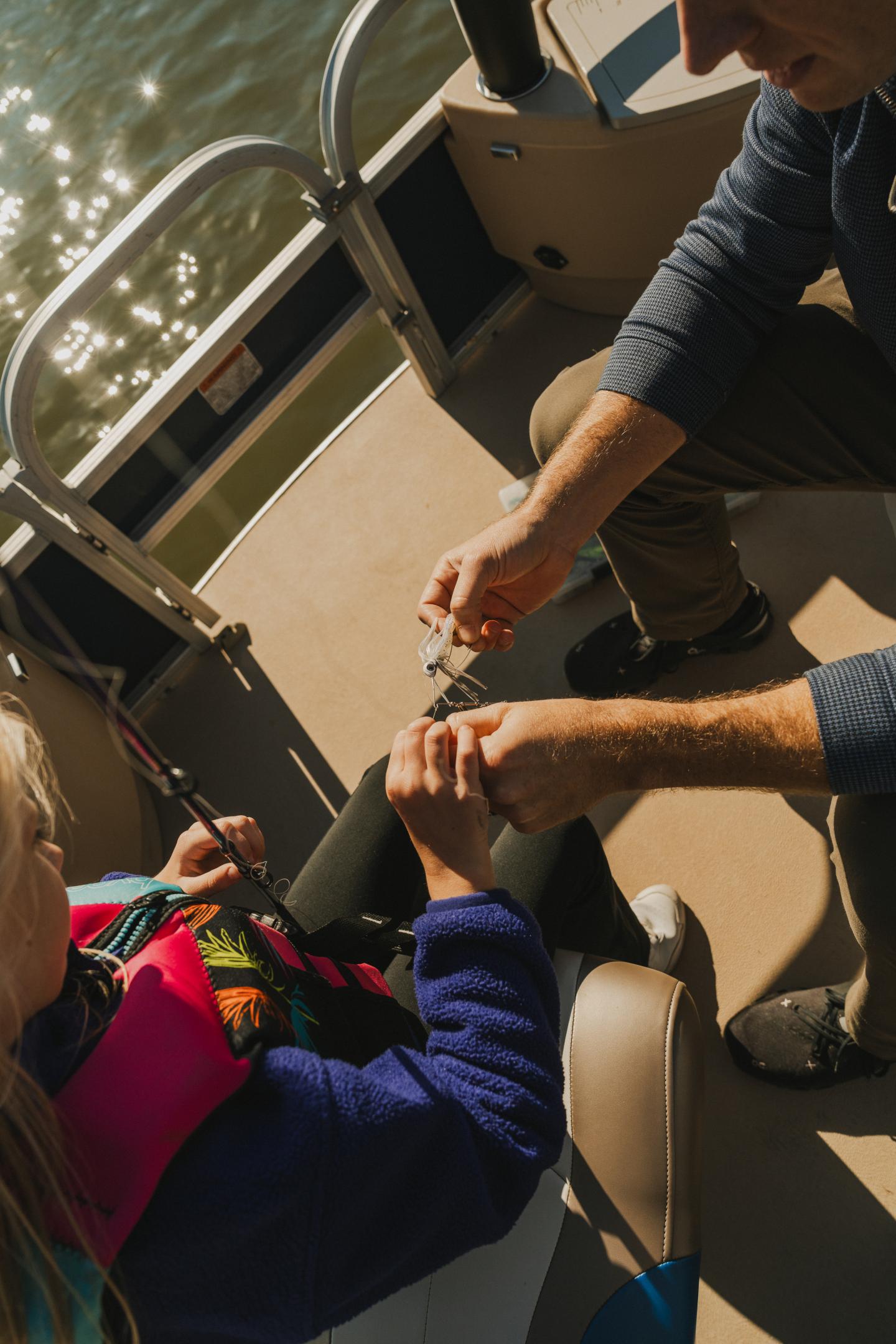 Two people in a boat, one tying a knot on fishing line, sunlight glinting off water.
