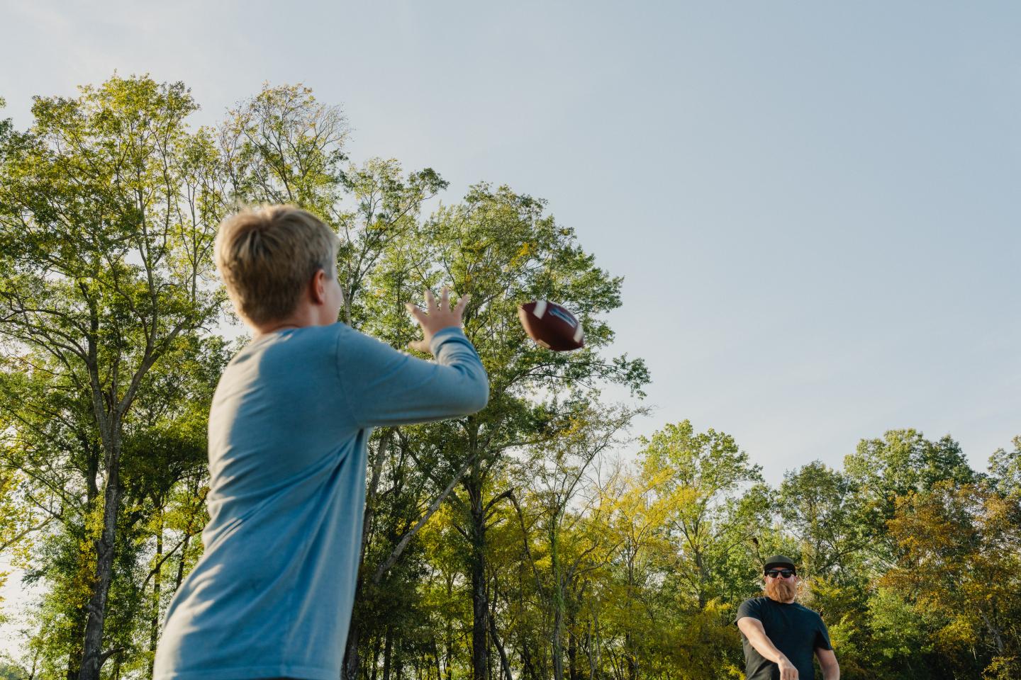 Boy throwing a football outdoors with trees and clear sky.