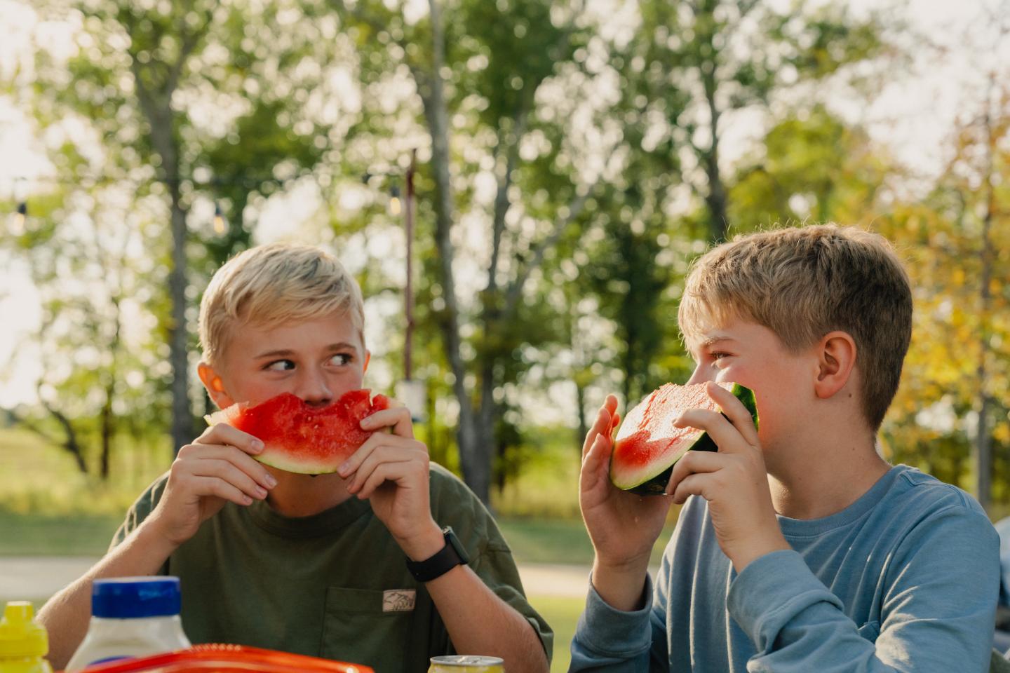 Two boys outdoors eating watermelon slices, smiling at each other.