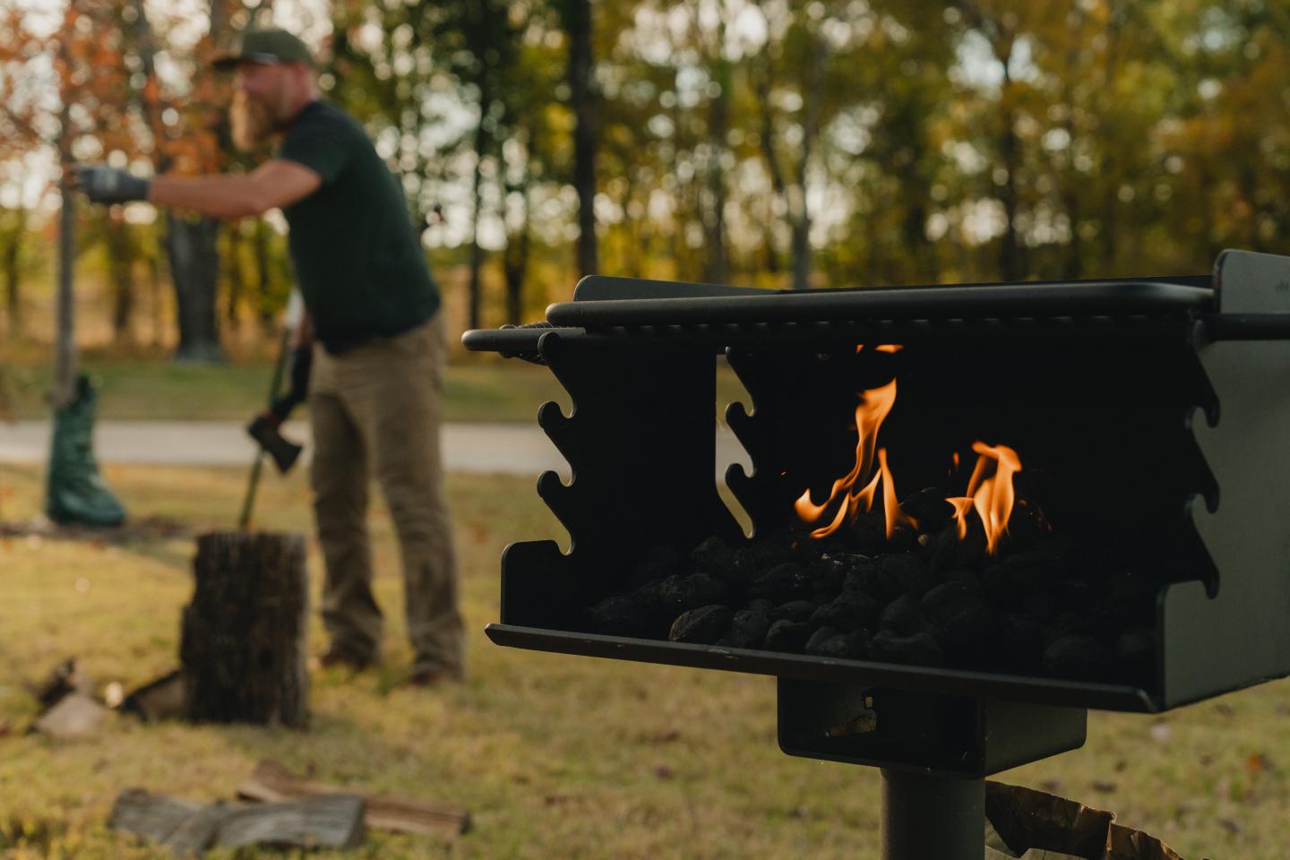 Outdoor grill with flames; man chopping wood nearby.