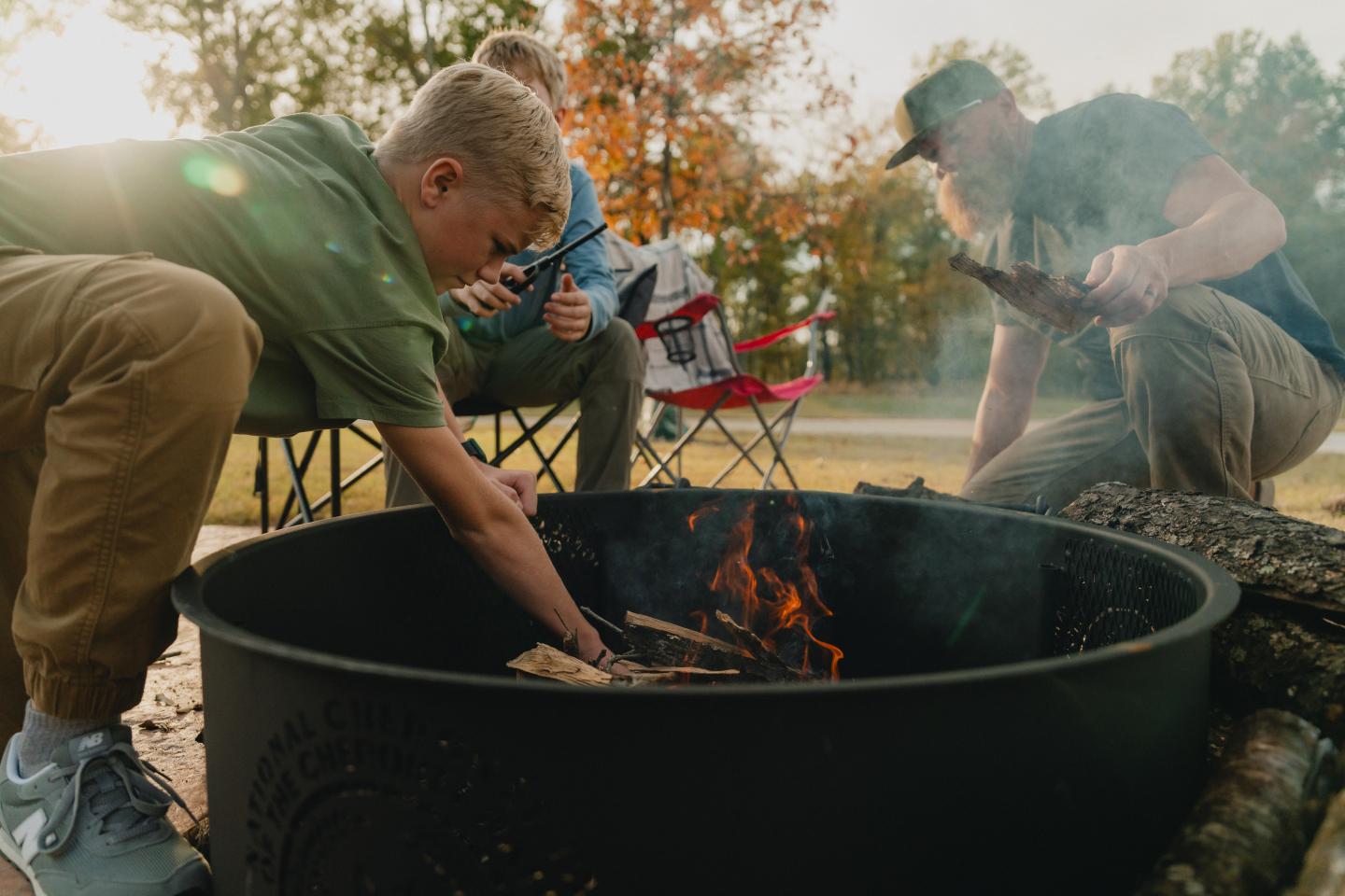 Three people sitting around a fire pit in a park, adding wood to the fire.