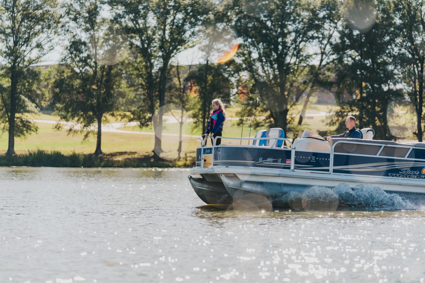 Pontoon boat on a lake with people, surrounded by trees.