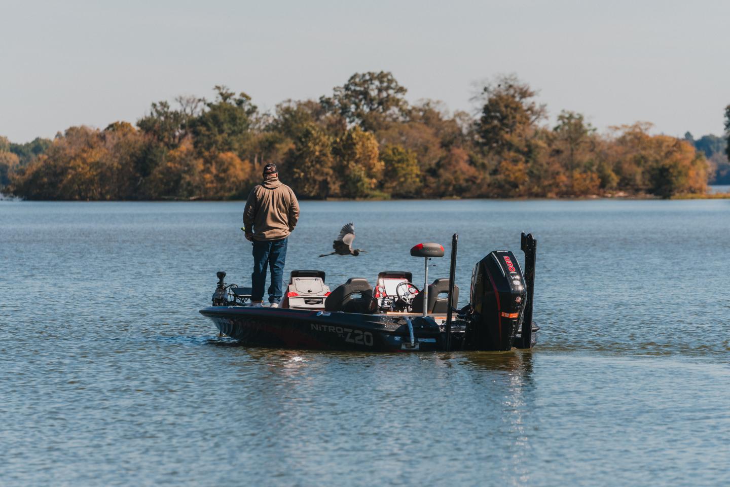 Man standing on a boat fishing in a calm lake with autumn trees in the background.