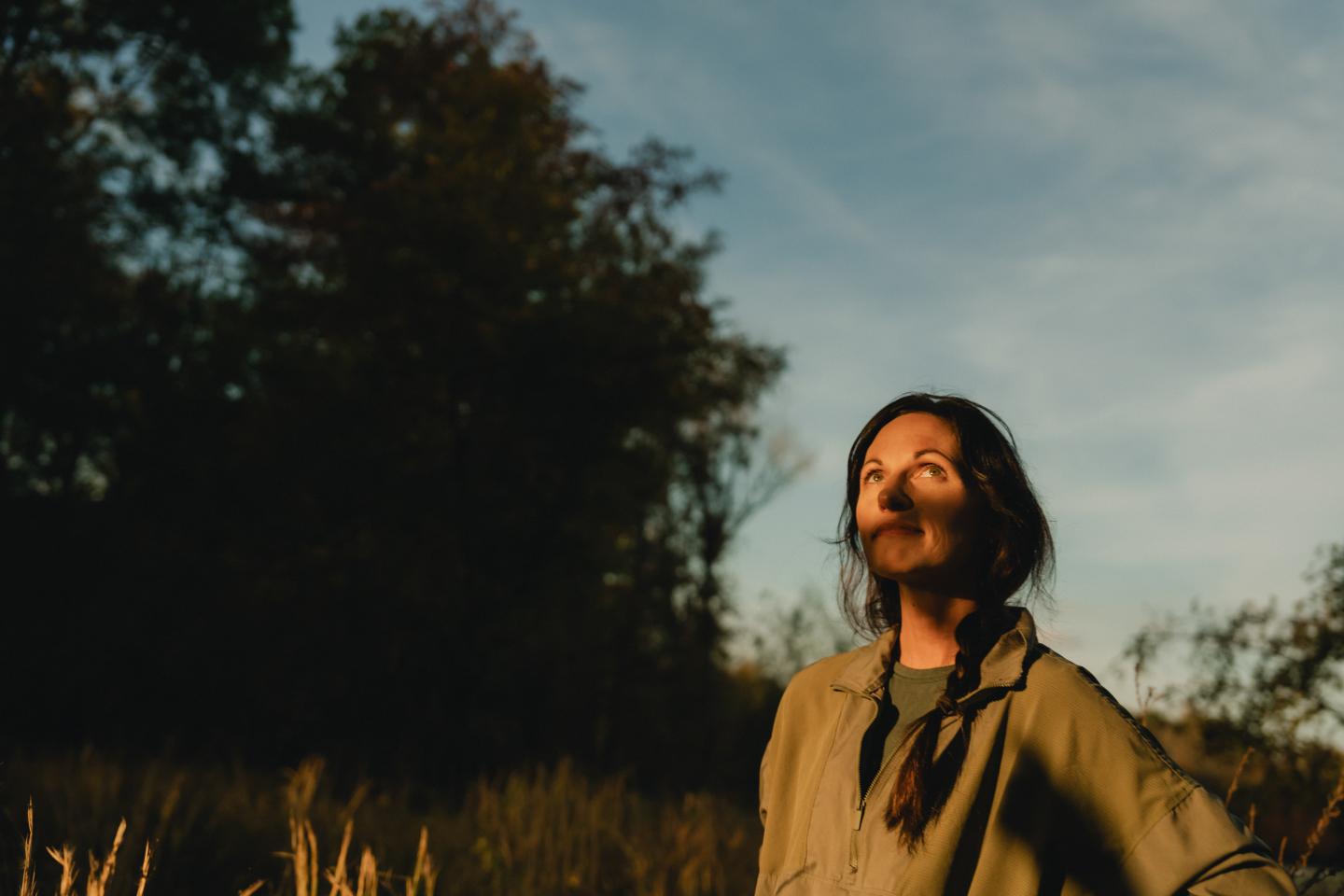 Woman in jacket smiles while looking at the sky in a sunlit field.