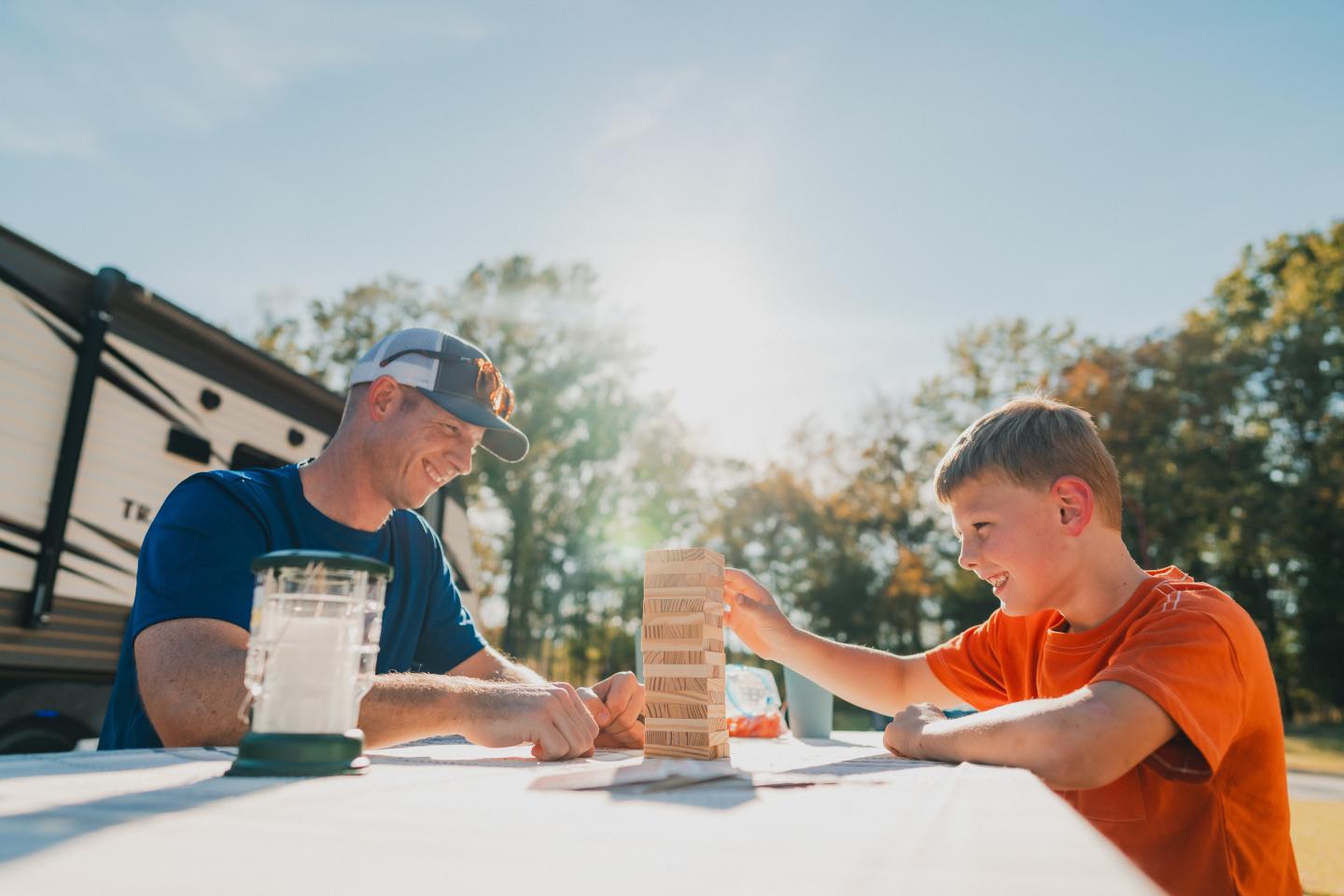 Father and son play Jenga at a picnic table outdoors, sunlight shining.