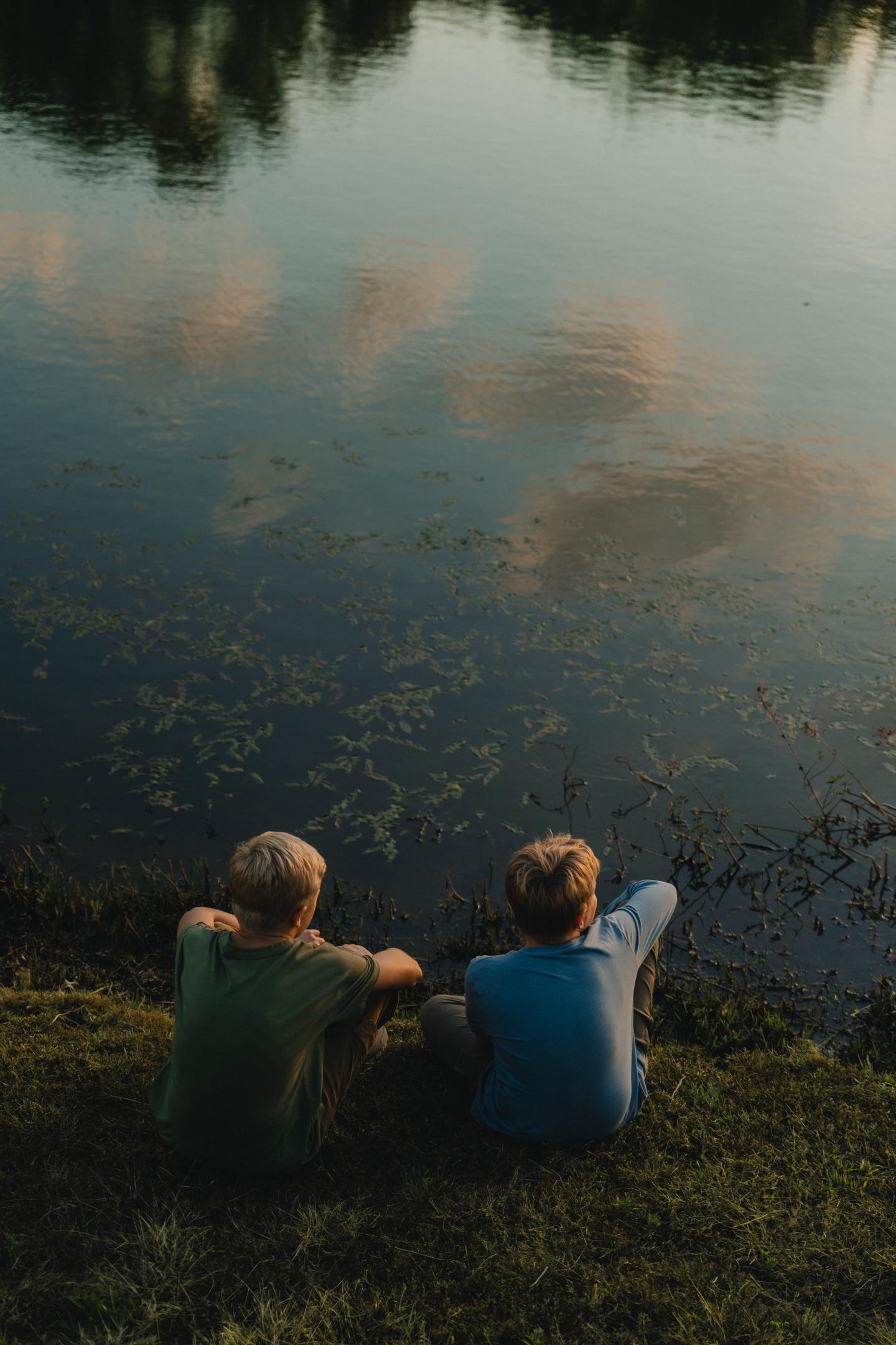 Two boys sit by a reflective, calm lake at sunset.