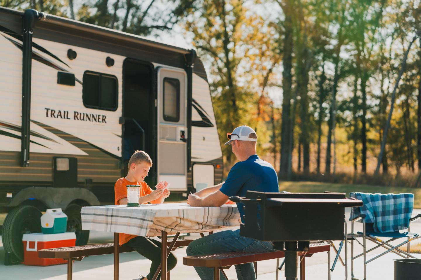 Father and child sitting at a table outside an RV, surrounded by trees.