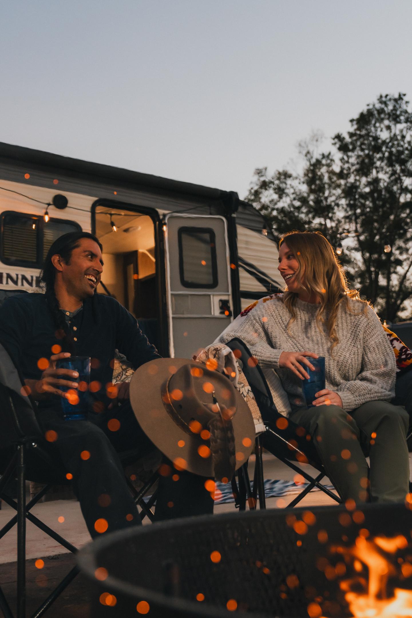 Campers sitting by a fire pit, trailer in background, dusk setting.