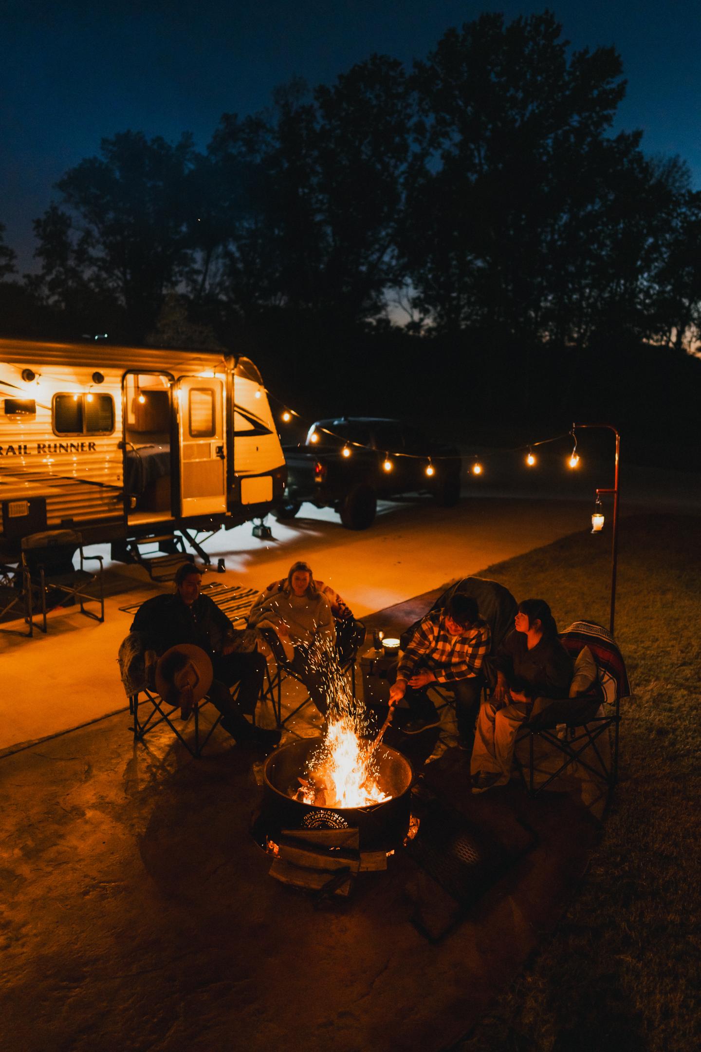 Camping scene at night with people around a fire, RV and string lights nearby.