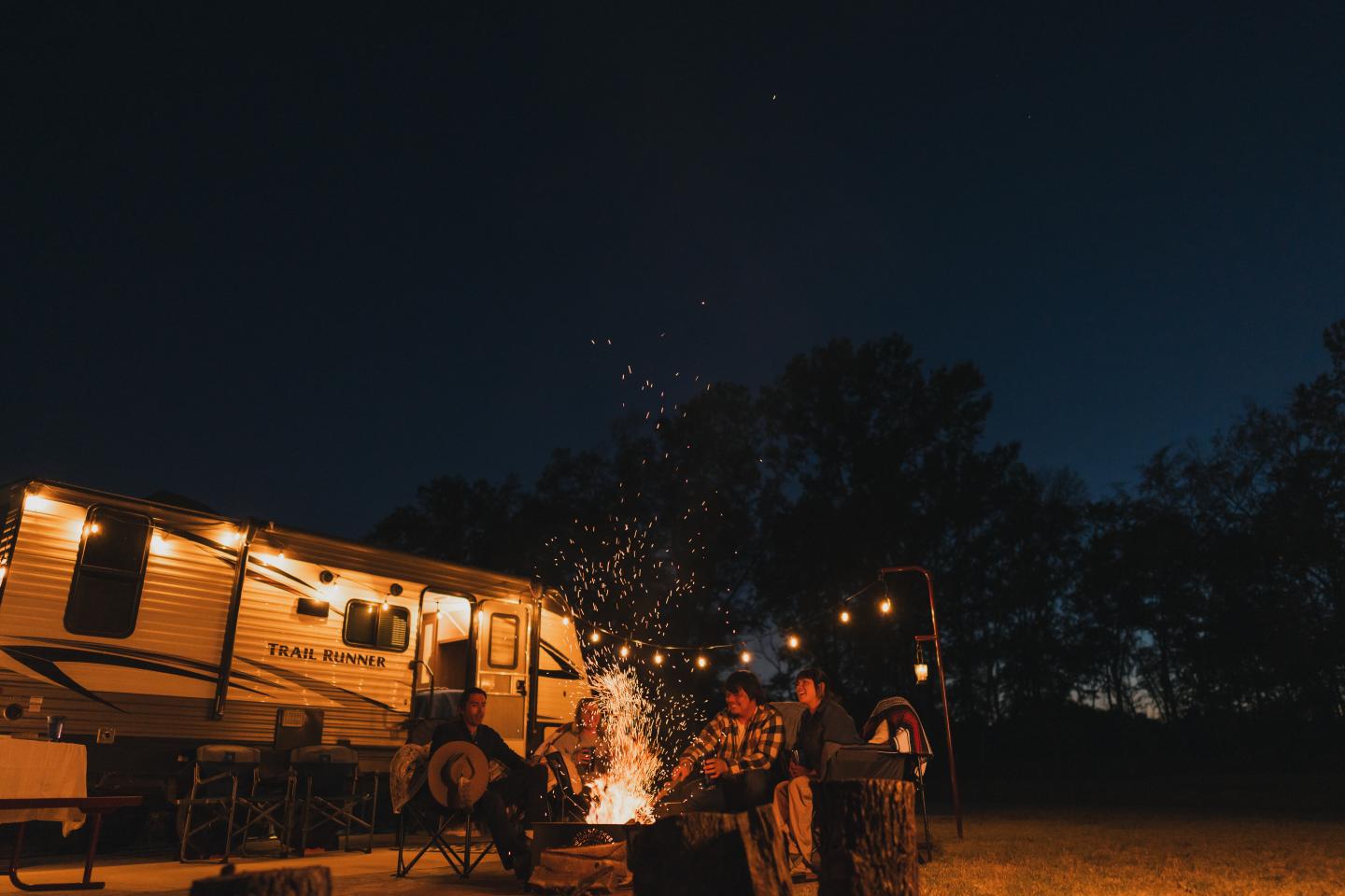 Campsite with RV, lit fire pit, and people seated under a starry night sky.