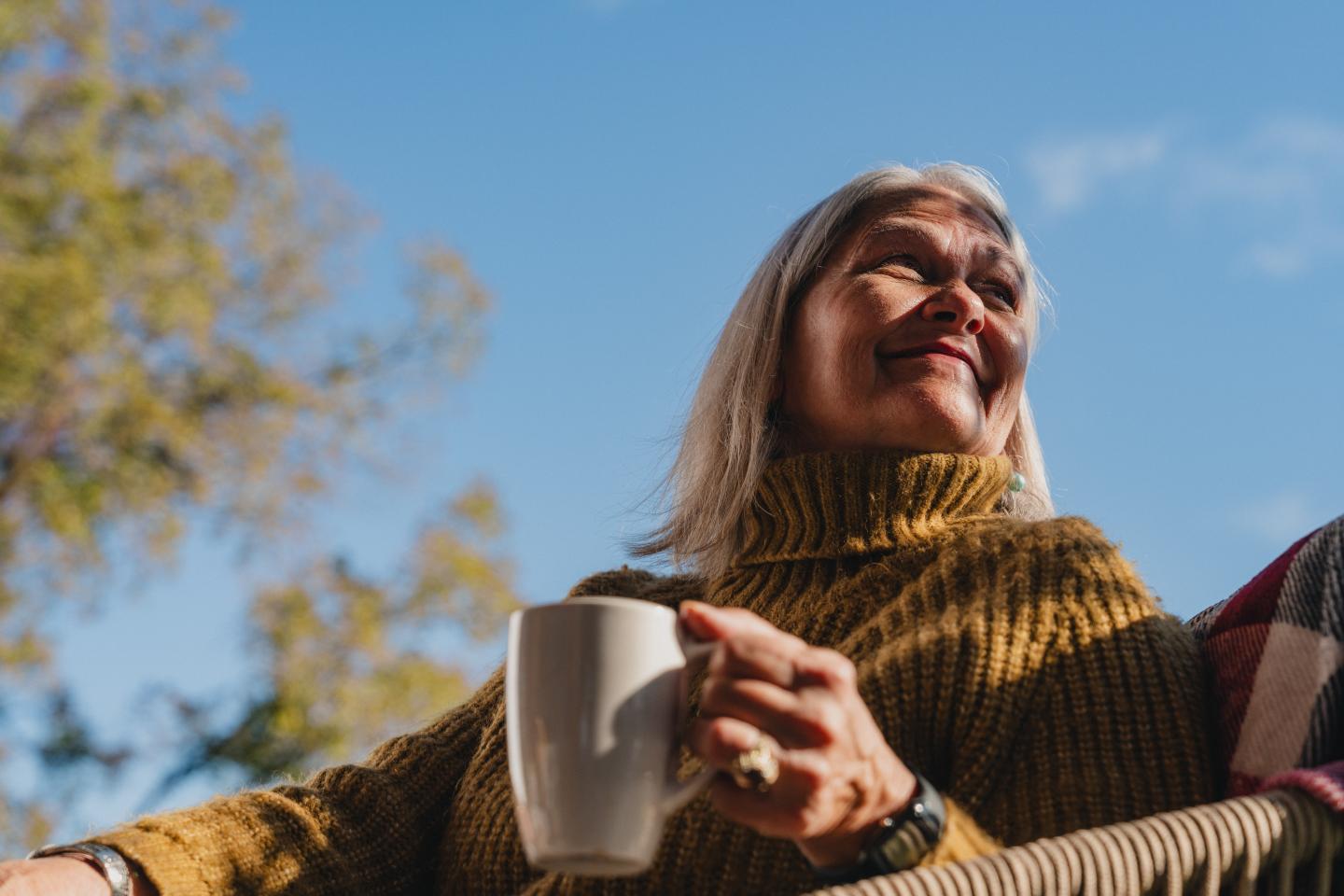 Smiling woman holding a mug, sitting outdoors with a blue sky background.