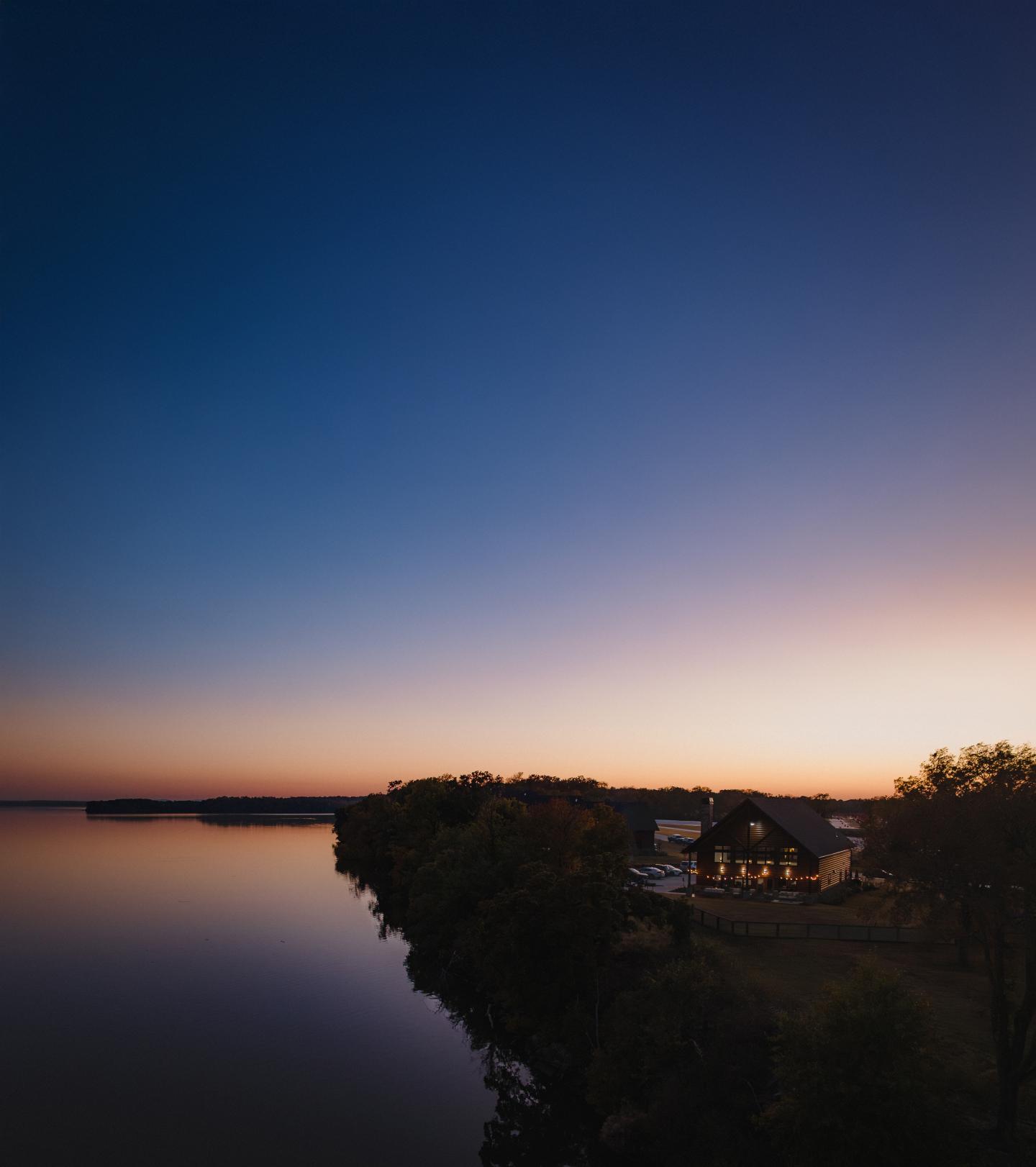 Sunset over a calm lake with a cabin and trees on the shoreline.