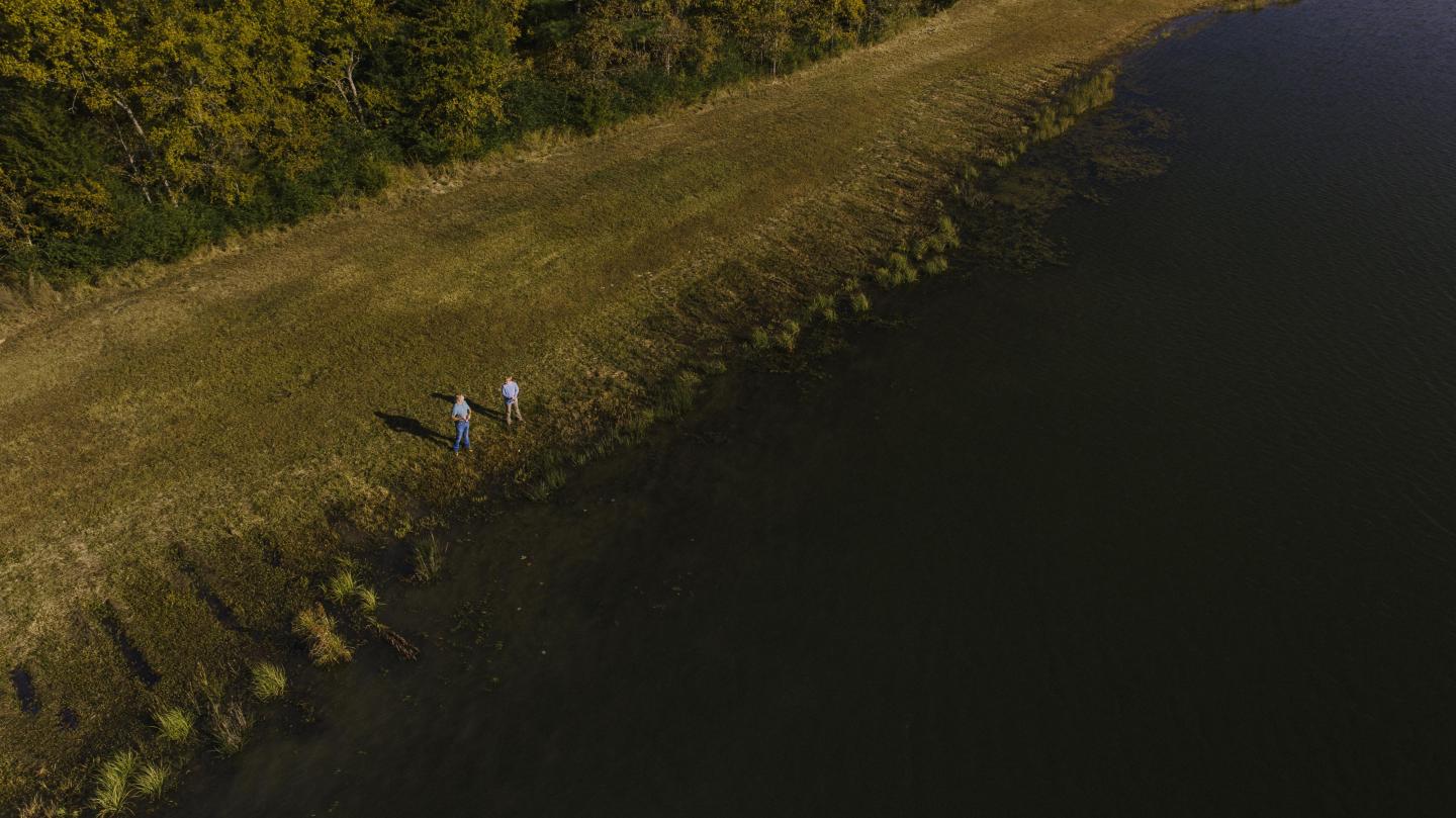 Two people walking along a grassy path beside a large dark lake, surrounded by trees.