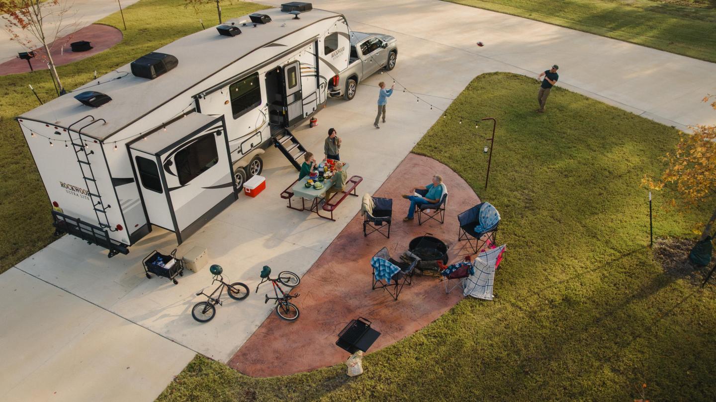 RV parked in a campground with people relaxing outside, bikes nearby.