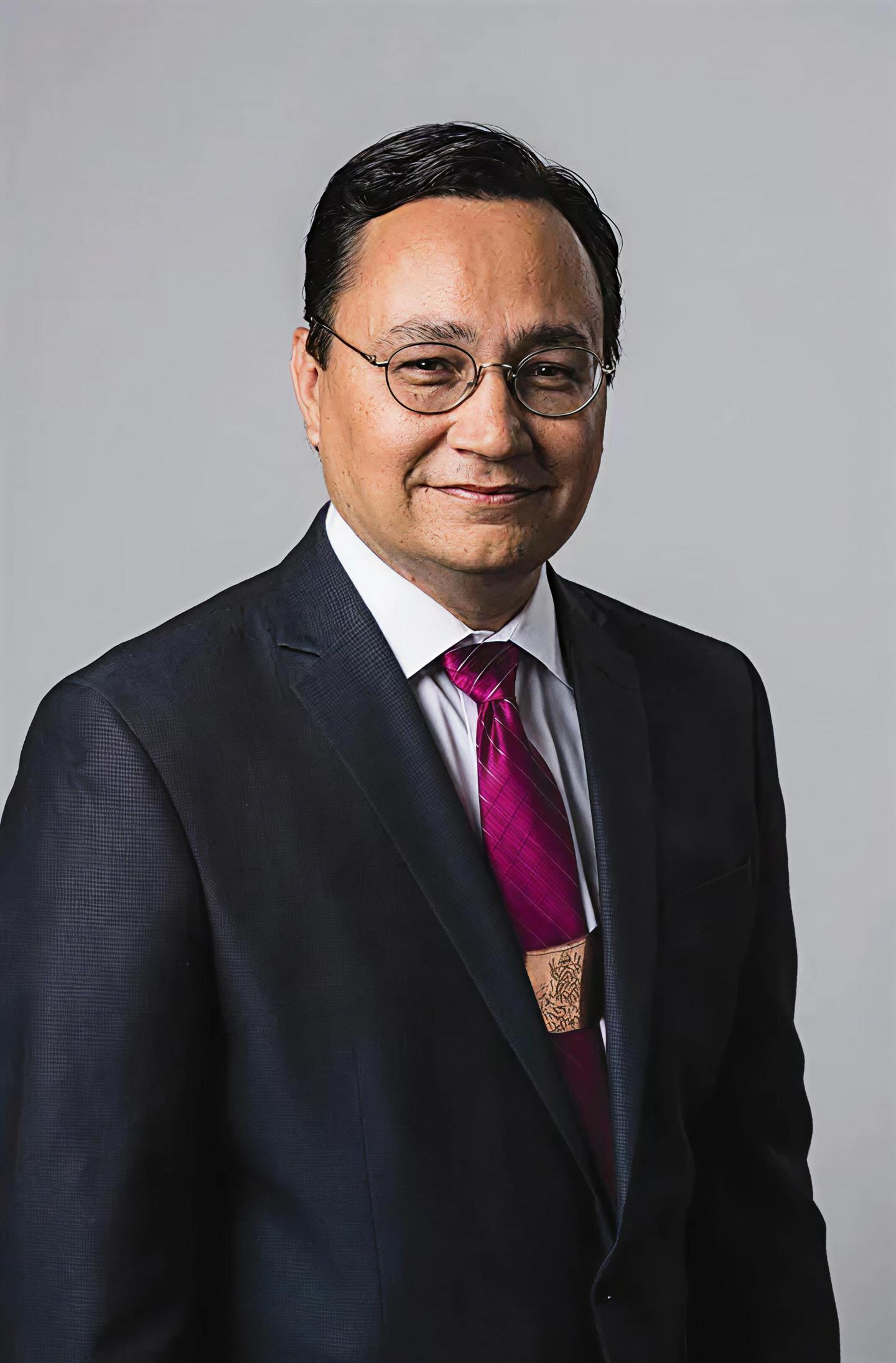 Man in a suit with glasses and a magenta tie, smiling slightly.