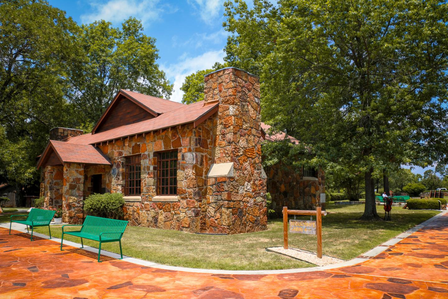 Stone house with red roof, green benches, surrounded by trees under blue sky.
