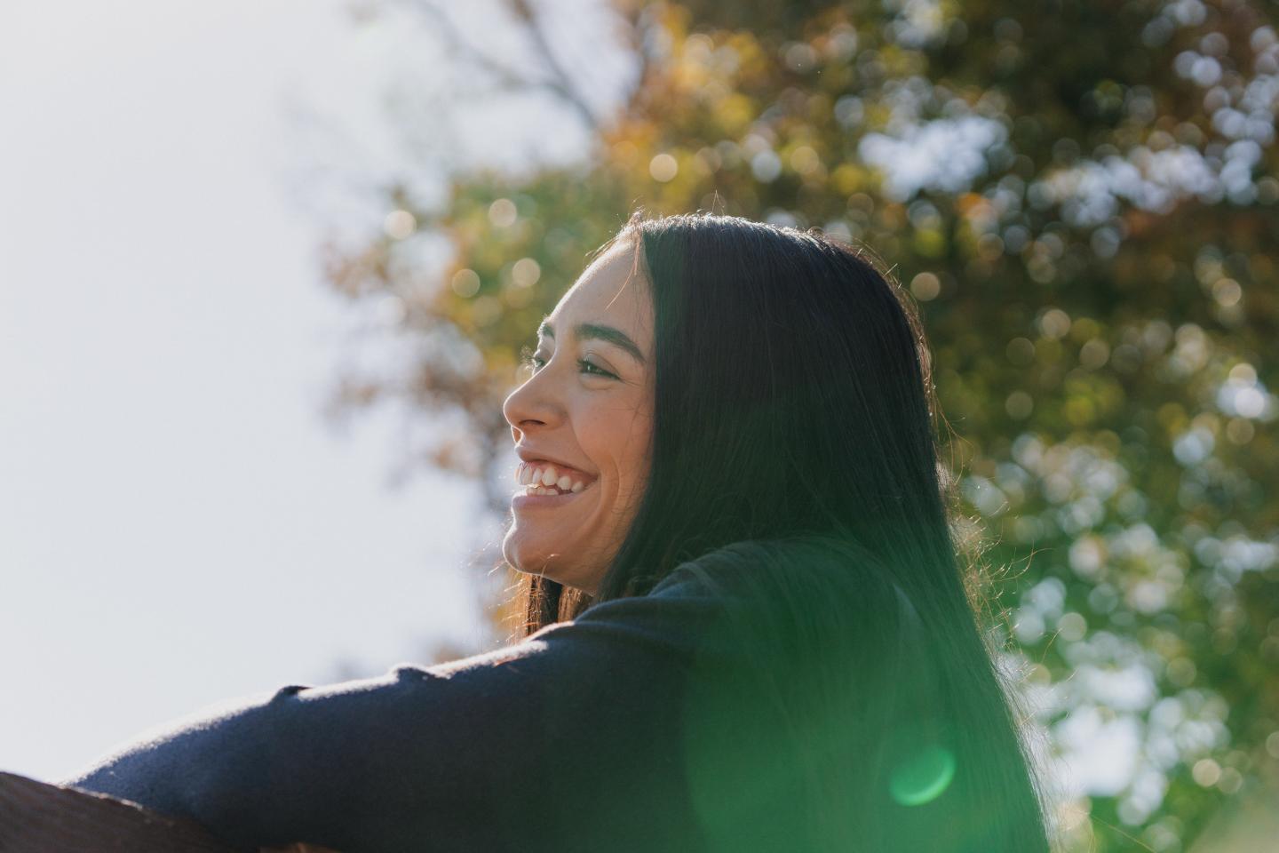 Smiling woman outdoors, sunlight filtering through trees in background.