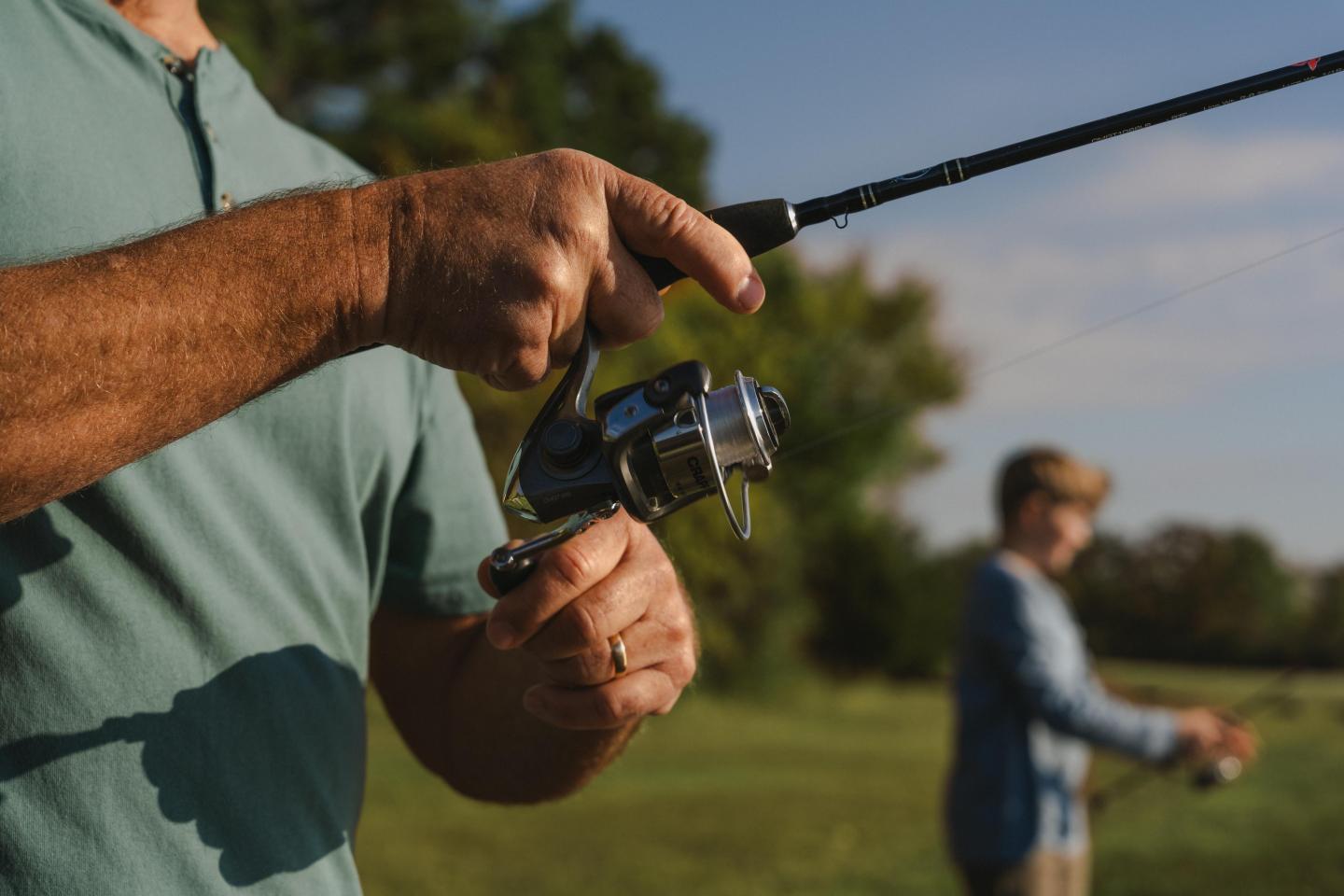 Two people fishing in a grassy area, one foreground, another blurred in background.