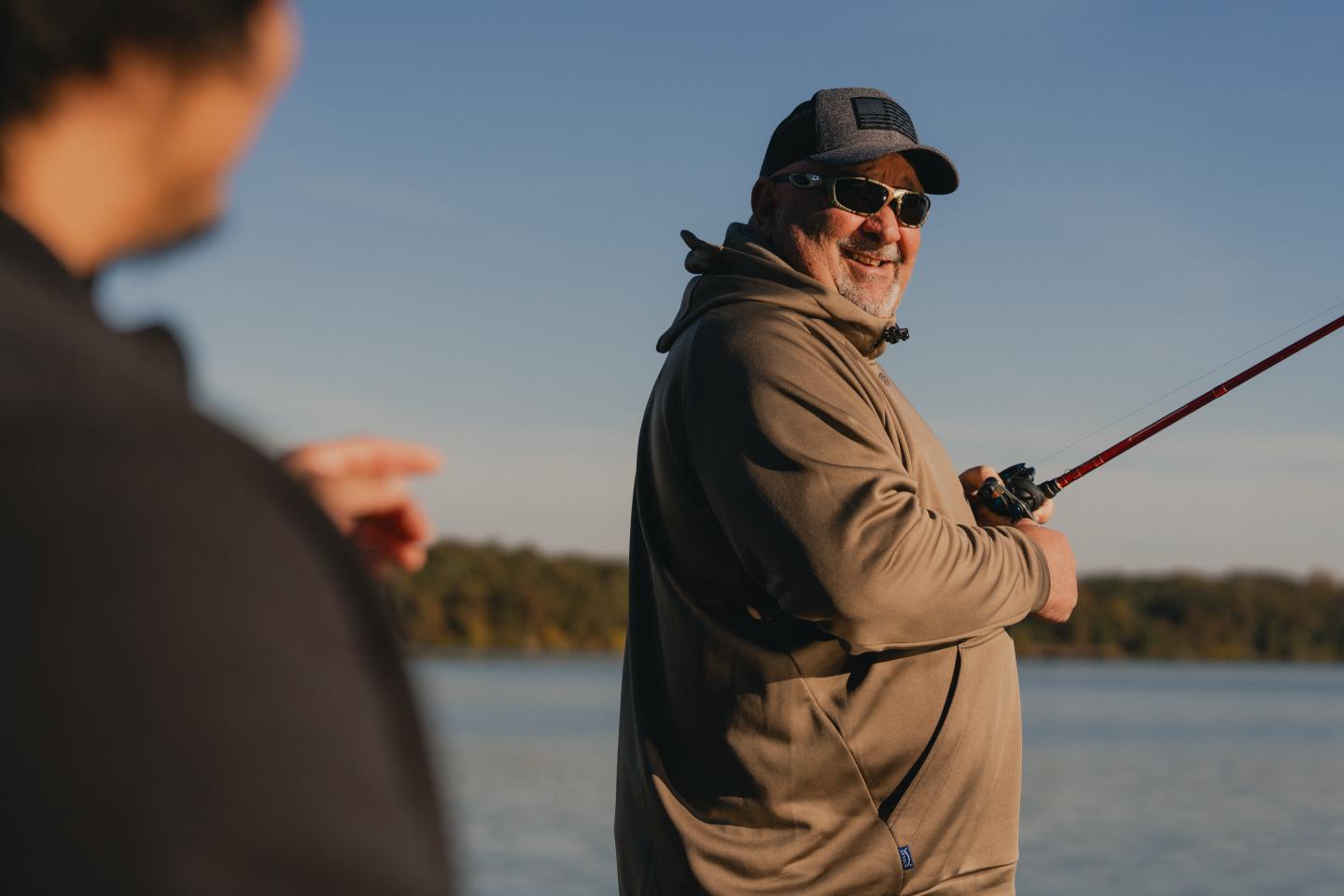 Man fishing by a lake, smiling, wearing sunglasses and a cap.
