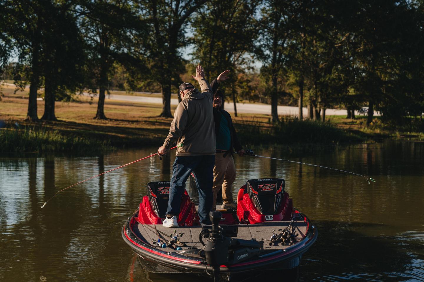 Two people fishing on a boat, surrounded by trees and calm water.