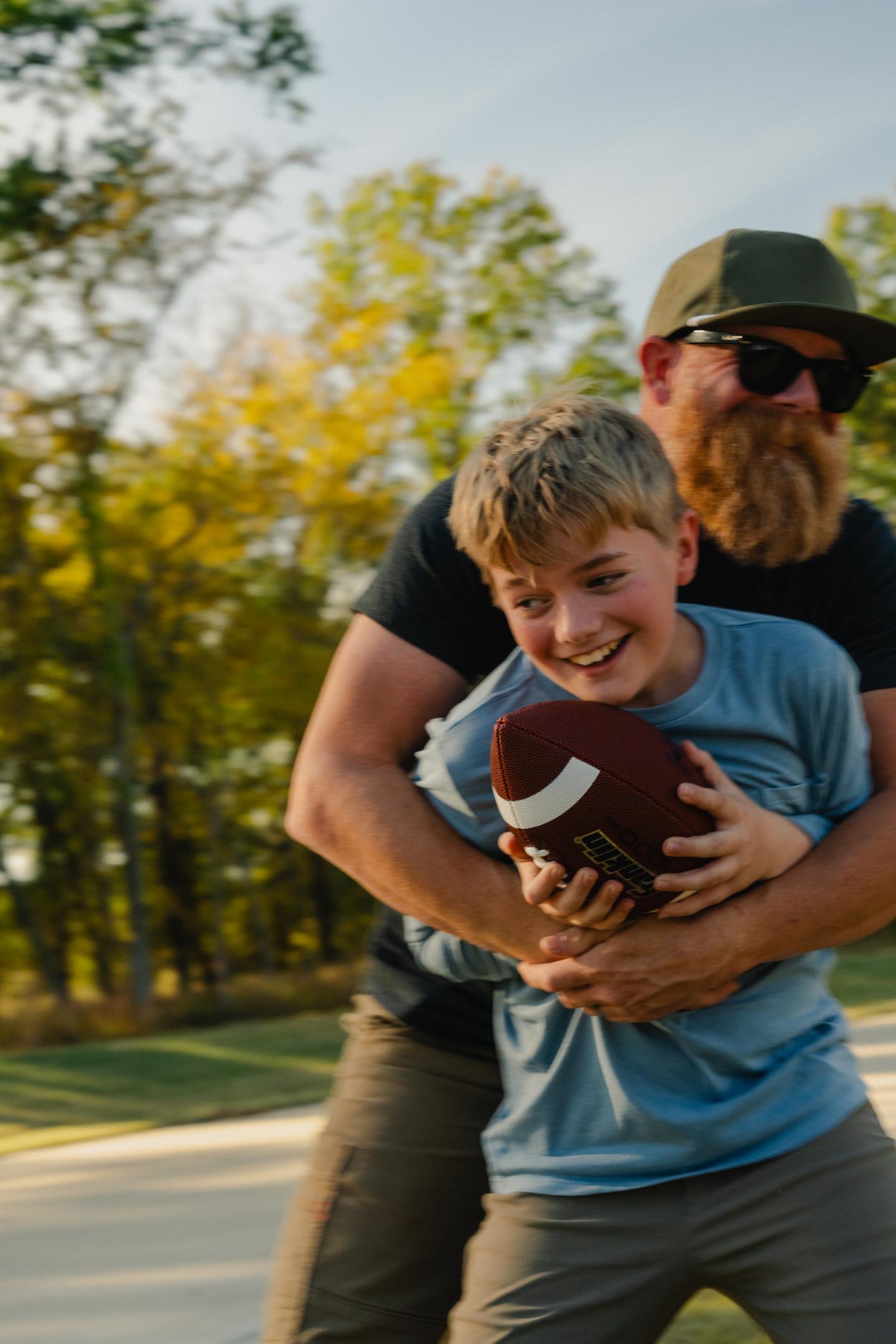 Adult playfully hugs boy holding football, outdoors, sunny day.