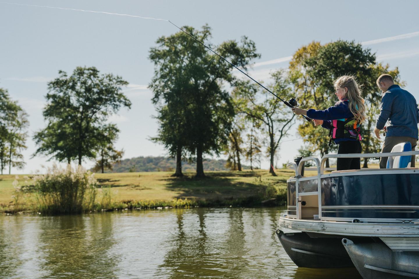 Girl and boy fishing from a boat on a sunny lake with trees in the background.