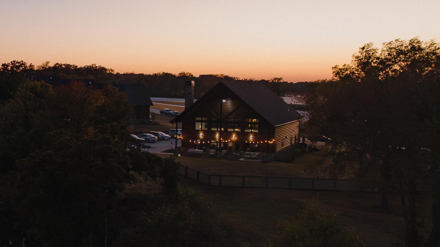 Cozy cabin with warm lights at sunset, surrounded by trees and a fenced yard.