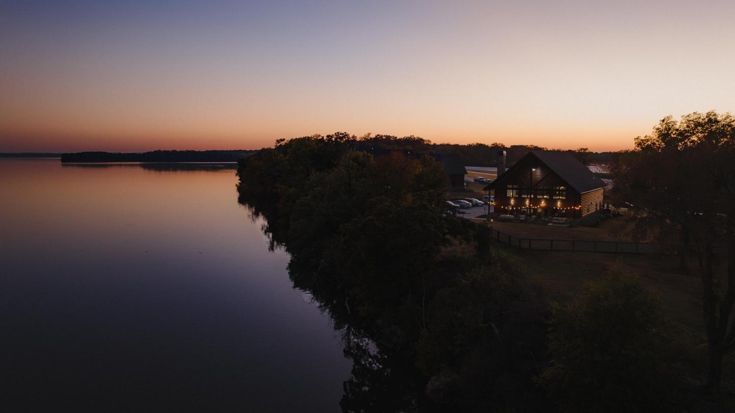 Lakeside house at sunset, warm lights on, serene water and twilight sky.